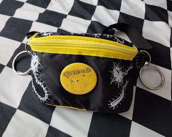 May include: A black bum bag with a bright yellow zip and trim. The bag features a white graphic print of spiked maces and chains. A circular yellow pin is attached to the front, displaying a line drawing of Pikachu and the text CHUDOS.