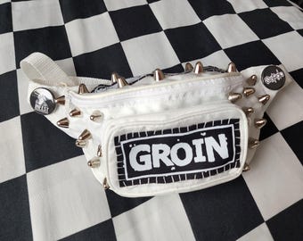 May include: A white bum bag featuring silver metal spikes along the top and sides. The front pocket displays a rectangular black and white patch with the text GROIN. Two circular pinback buttons are attached, one reading INFEST and one Sordo.