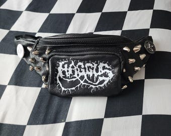 May include: A black bum bag with a textured surface, featuring silver-toned metal spikes on the sides and a front pocket with a white patch displaying the text HAGGUS in a stylised, jagged font. A circular pin is attached to the side.