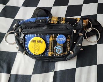 May include: A black bum bag featuring blue and yellow plaid fabric patches, silver metal spikes, and two large silver rings on the sides. It is decorated with three circular pins: a yellow one with a cartoon character, a blue one, and a black one.