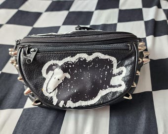 May include: A black leather bum bag featuring a white and black sheep graphic on the front pocket. The bag has a zippered main compartment and a smaller front zippered pocket, accented with silver-toned metal spikes along both side edges.