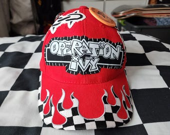 May include: A red baseball cap featuring a black and white embroidered patch with the text Operation Ivy. The brim displays a black and white checkered pattern overlaid with red flame-shaped embroidery. A circular orange pin is attached to the top.