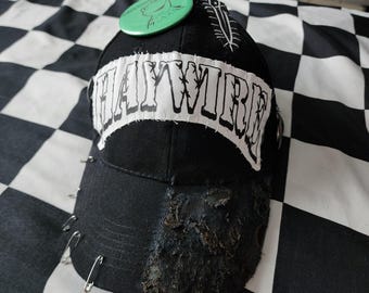 May include: A black baseball cap featuring a white fabric patch with the text HAYWIRE in black lettering. The brim is distressed with heavy fraying and multiple silver safety pins attached to the left side. A green circular pin sits on top.