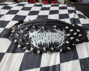 May include: A black bum bag featuring a series of silver-toned metal spikes arranged along the top and bottom edges. The centre of the bag displays a white graphic print with intricate, jagged, and stylised lettering in a heavy metal aesthetic.