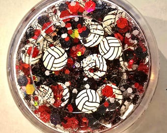 May include: A clear round container filled with black, red, and white glitter. The glitter is shaped like volleyballs and stars.