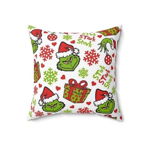 May include: Square white pillow with a repeating pattern of a green character wearing a Santa hat, red and green snowflakes, red hearts, and wrapped gifts. The words "Stink Stunk" are also printed on the pillow.