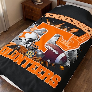 Puede incluir: Manta negra con las palabras "Tennessee" y "Volunteers" en naranja. El diseño incluye una gran "T" naranja, un casco de fútbol americano, un balón de fútbol y una mano de espuma. La manta está sobre una cama.