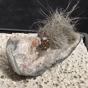May include: A natural mineral specimen featuring a geode with a cluster of needle-like crystals. The geode has a white and gray exterior with a cavity filled with small, sparkling crystals and a cluster of metallic-looking crystals.