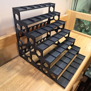 May include: A black tiered display stand featuring five levels of shelving. The side panels are constructed with a geometric honeycomb pattern. Each shelf is flat and rectangular, designed to hold small items in an organized, stepped arrangement.