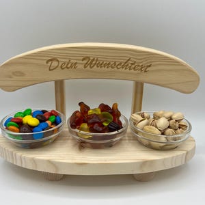 Könnte beinhalten: Holz-Snack-Ständer mit drei Glasschalen. Die Schalen enthalten bunte Süßigkeiten, Gummibärchen und Pistazien. Der Ständer hat eine gebogene Oberseite mit dem eingravierten Text "Dein Wunschtext".