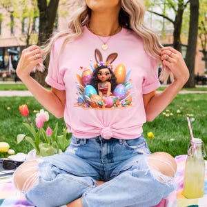 May include: Pink t-shirt featuring a cartoon character with bunny ears surrounded by Easter eggs. The design includes a character with dark hair and a red and white top. The shirt is tied at the waist and paired with ripped blue jeans.