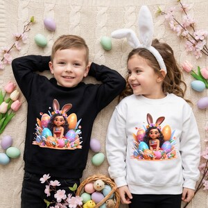 May include: Two children wearing Easter-themed sweatshirts. One child wears a black sweatshirt, the other a white one. Both sweatshirts feature a graphic of a girl with bunny ears and colorful Easter eggs. The children are smiling.