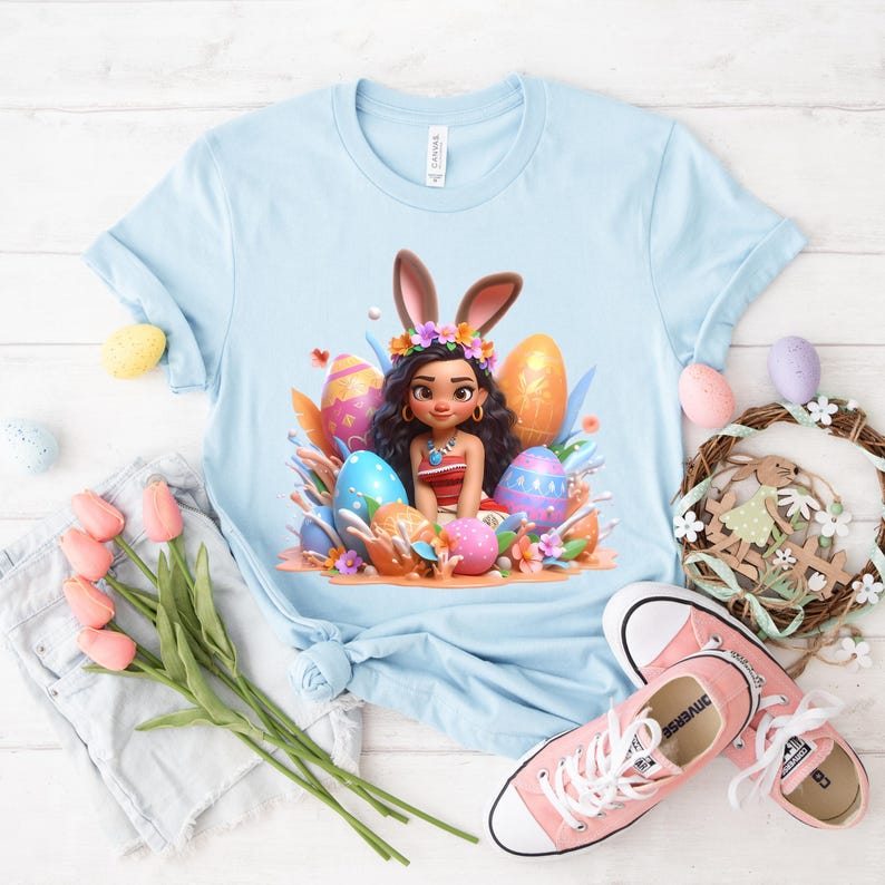 May include: Light blue t-shirt featuring a cartoon illustration of a girl with bunny ears surrounded by Easter eggs. The eggs are decorated with various patterns and colors, including pink, blue, and gold. The shirt is paired with pink sneakers and a denim skirt.