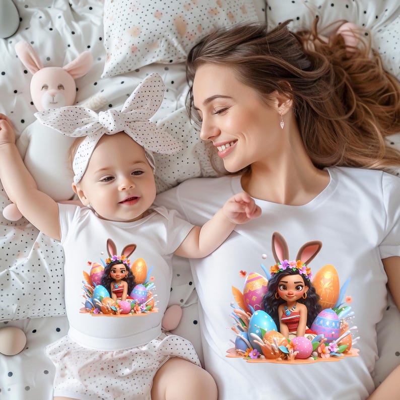 May include: A mother and baby wearing matching white t-shirts with an Easter-themed graphic. The graphic features a cartoon character with bunny ears surrounded by colorful Easter eggs and flowers. The baby wears a white bow headband.
