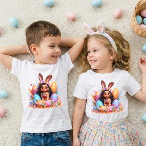 May include: Two children wearing white t-shirts with an Easter-themed graphic. The graphic features a cartoon character with bunny ears surrounded by colorful Easter eggs. The children are lying on a light-colored rug, with Easter eggs scattered around them.
