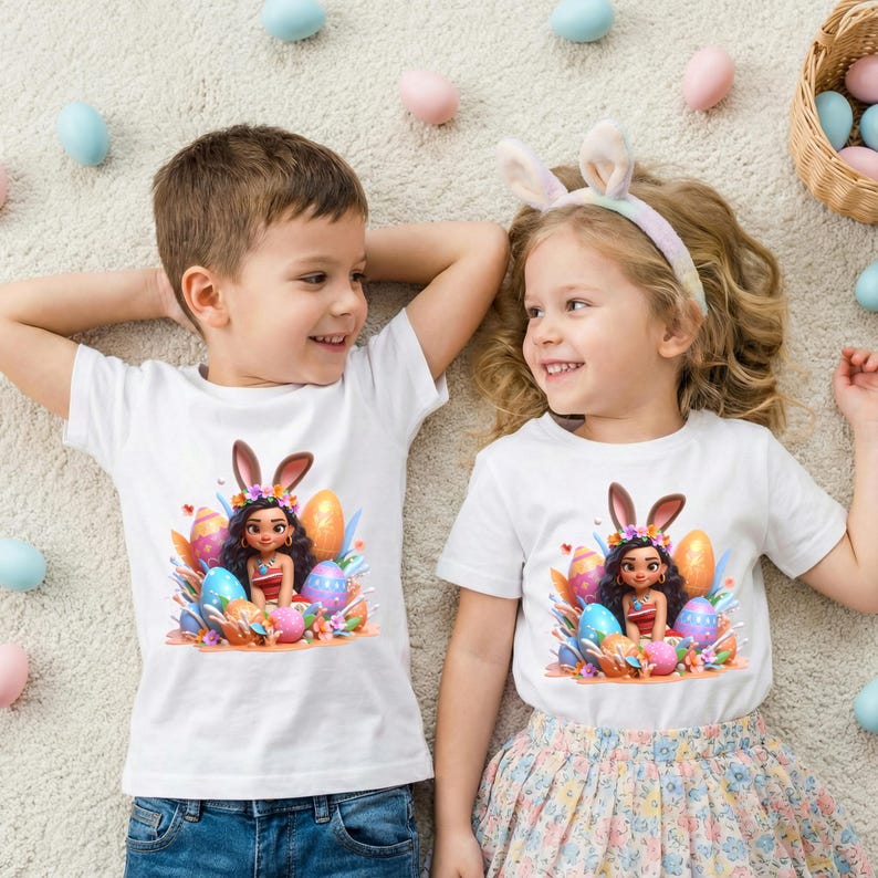 May include: Two children wearing white t-shirts with an Easter-themed graphic. The graphic features a cartoon character with bunny ears surrounded by colorful Easter eggs. The children are lying on a light-colored rug, with Easter eggs scattered around them.