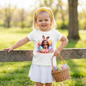 May include: A young child wearing a white t-shirt with an Easter-themed graphic, a yellow headband, and a white pleated skirt. The graphic features a cartoon character with bunny ears surrounded by colorful Easter eggs. The child is holding a wicker basket filled with decorated eggs.