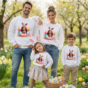 May include: A family wearing matching white sweatshirts with a colorful Easter-themed graphic. The graphic features a cartoon character with bunny ears surrounded by Easter eggs. The family is standing in a garden with a basket of Easter eggs.