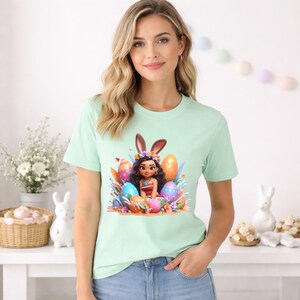 May include: A light green t-shirt featuring a colorful Easter-themed graphic. The graphic depicts a cartoon character with bunny ears, surrounded by decorated Easter eggs. The character is wearing a floral crown and a red and white outfit.