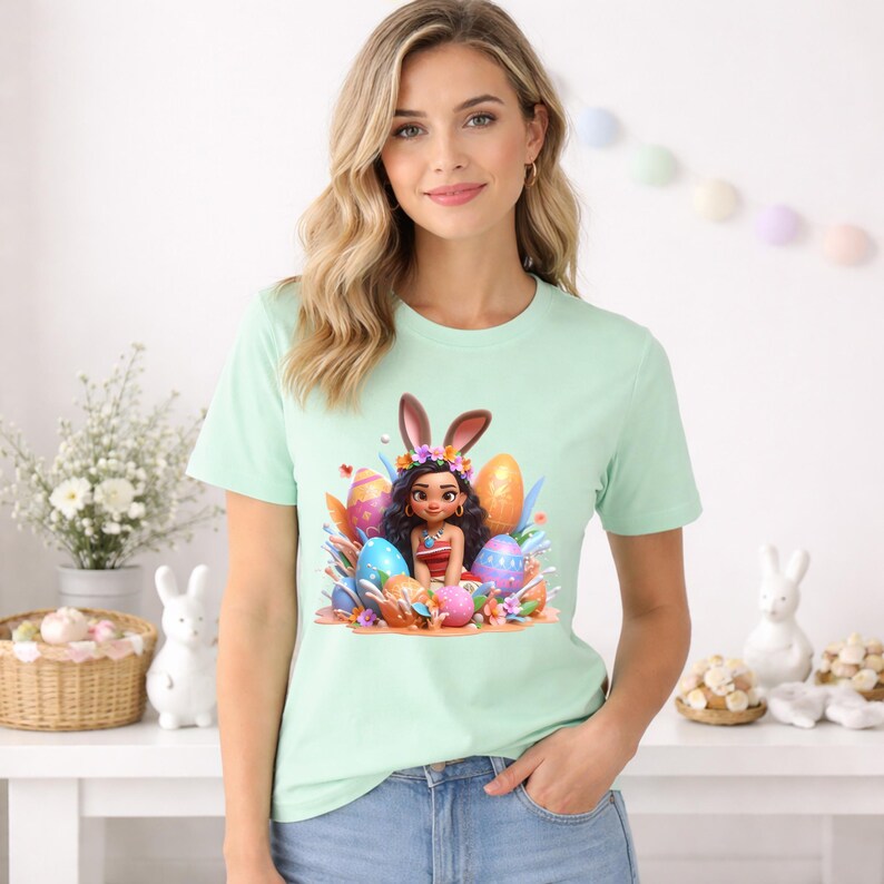 May include: A light green t-shirt featuring a colorful Easter-themed graphic. The graphic depicts a cartoon character with bunny ears, surrounded by decorated Easter eggs. The character is wearing a floral crown and a red and white outfit.