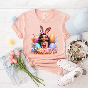 May include: A light peach-colored t-shirt featuring a cartoon illustration of a girl with bunny ears surrounded by Easter eggs. The image includes a white cap, a wreath, and a pair of white sneakers. A bouquet of pink tulips and a pair of denim shorts are also present.