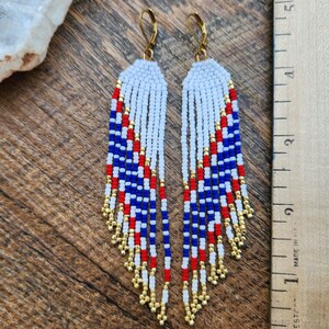 May include: A pair of long, beaded earrings with a red, white, and blue color scheme. The earrings are made with small, round beads and have a gold-colored wire loop at the top.