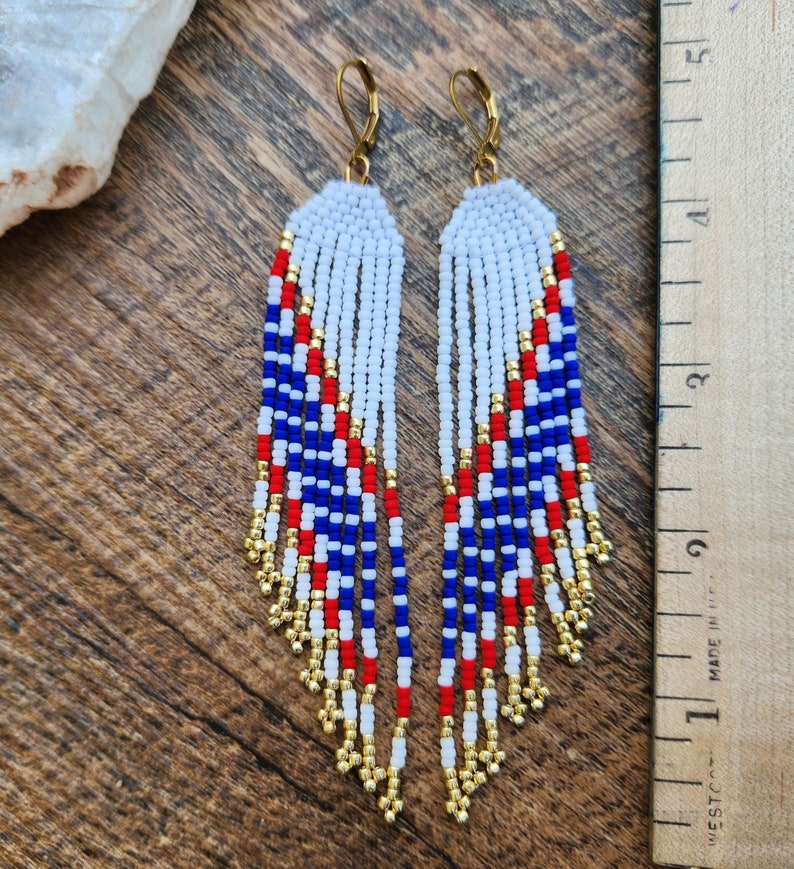 May include: A pair of long, beaded earrings with a red, white, and blue color scheme. The earrings are made with small, round beads and have a gold-colored wire loop at the top.