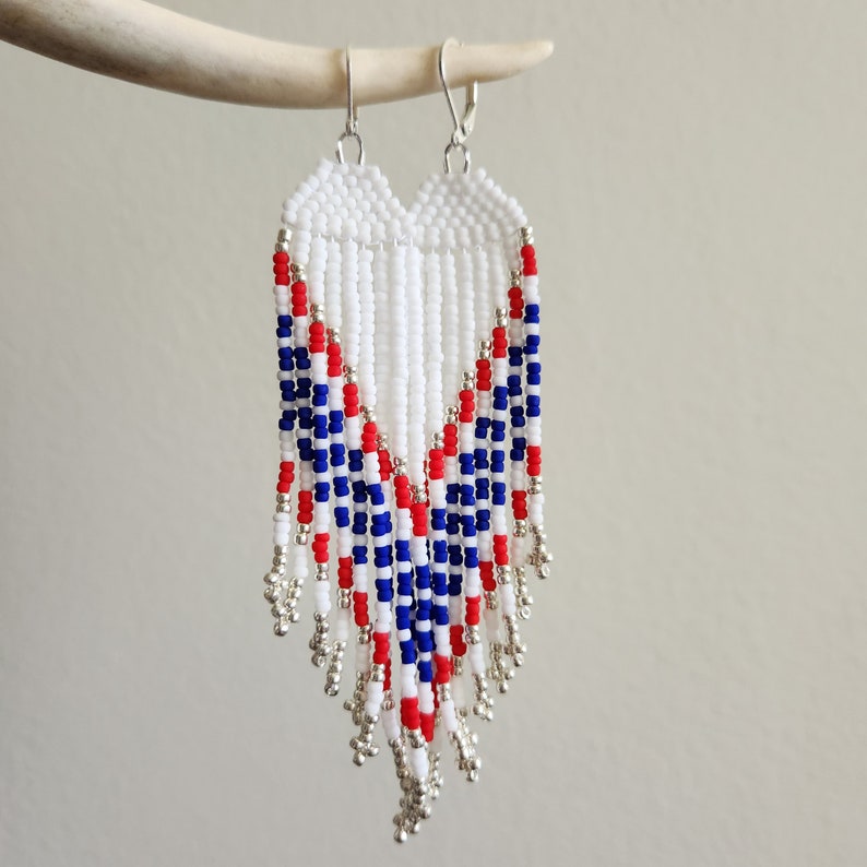 May include: A pair of white, red, and blue beaded earrings with silver accents. The earrings are in the shape of a teardrop and have a fringe design.