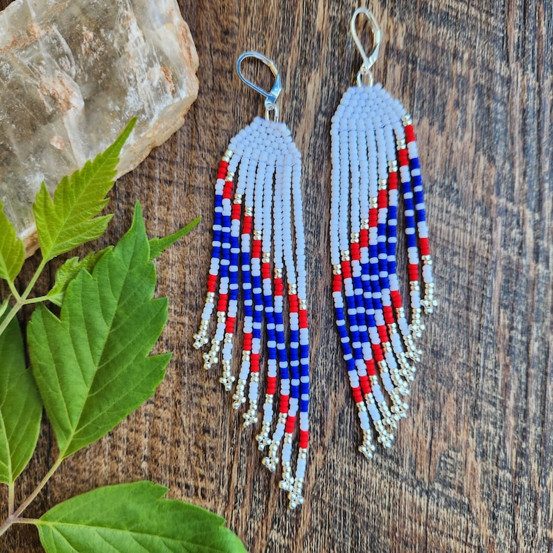 May include: A pair of long, beaded earrings with a white, red, and blue color scheme. The earrings are made with small, round beads and have a fringe-like design.