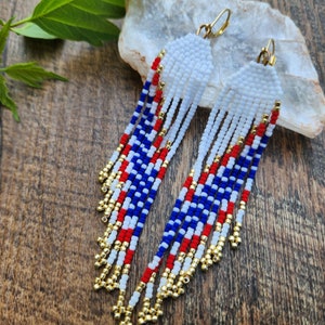 May include: A pair of long, beaded earrings with a red, white, and blue color scheme. The earrings are made with small, round beads and have a gold-colored wire.