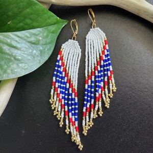 May include: A pair of long, beaded earrings with a red, white, and blue color scheme. The earrings are made of small, round beads and have a fringe-like design.