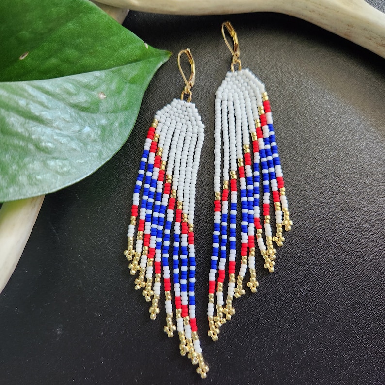 May include: A pair of long, beaded earrings with a red, white, and blue color scheme. The earrings are made of small, round beads and have a fringe-like design.