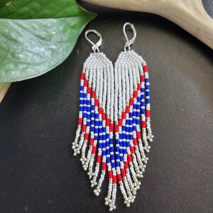 May include: A pair of long, beaded earrings with a white, red, and blue color scheme. The earrings are made with small, round beads and have a fringe-like design.
