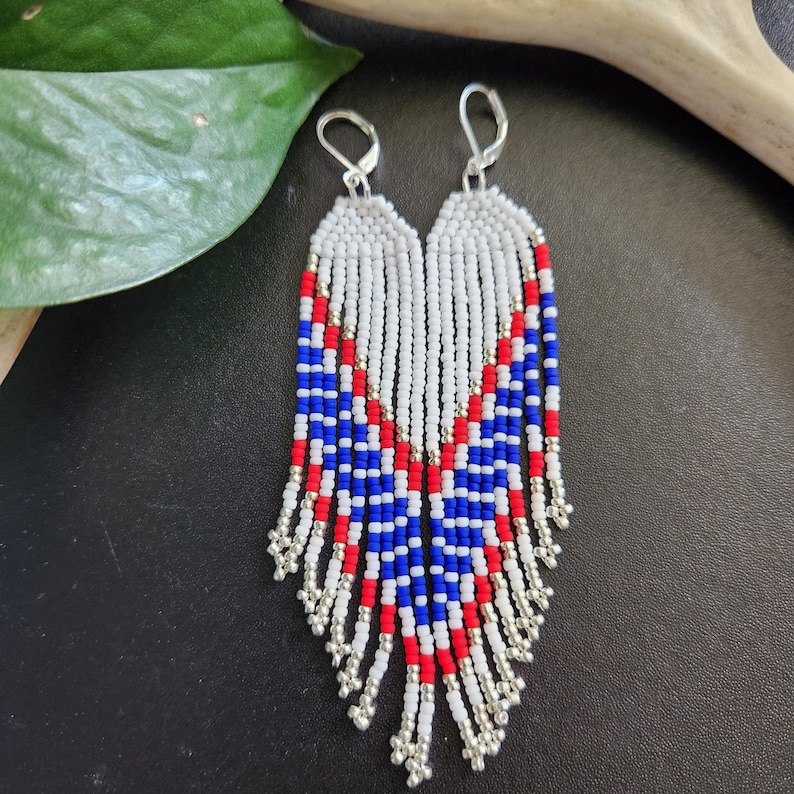 May include: A pair of long, beaded earrings with a white, red, and blue color scheme. The earrings are made with small, round beads and have a fringe-like design.