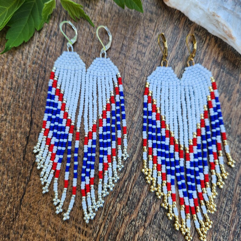 May include: A pair of long, beaded earrings with a white, red, blue, and gold color scheme. The earrings are made of small beads and have a fringe-like design.