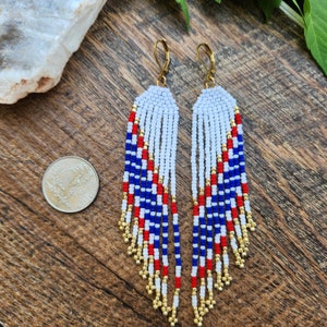 May include: A pair of long, beaded earrings with a red, white, and blue color scheme. The earrings are made with small, round beads and have a fringe-like design. The earrings are hanging from gold hooks.
