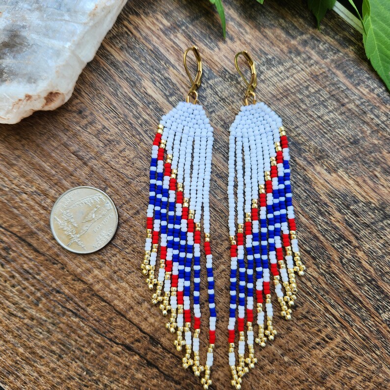 May include: A pair of long, beaded earrings with a red, white, and blue color scheme. The earrings are made with small, round beads and have a fringe-like design. The earrings are hanging from gold hooks.