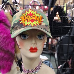 Puede incluir: Una cabeza de maniquí con una gorra de camionero de camuflaje verde con un parche amarillo y naranja que dice "KABOOM!". La gorra tiene una malla negra en la parte posterior y una visera blanca. El maniquí lleva un vestido azul y blanco y un collar de pedrería multicolor.