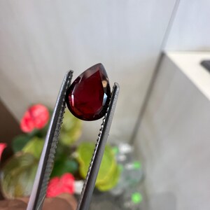 Può includere: Una pietra preziosa rosso scuro a forma di pera tenuta da una pinzetta di metallo. La pietra presenta un taglio sfaccettato che riflette la luce, mostrando una ricca tonalità cremisi scuro in tutto il suo corpo traslucido. La pinzetta ha una presa dentellata.