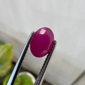 May include: A vibrant, oval-cut magenta gemstone held securely by a pair of silver-toned metal tweezers. The stone features a faceted surface that reflects light, showcasing a deep, saturated pinkish-red color throughout its polished body.