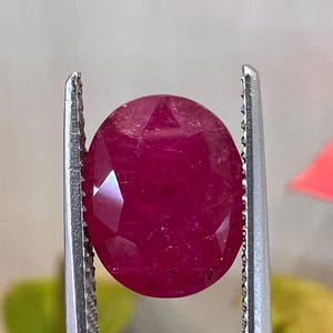 Könnte beinhalten: Ein oval geschliffener, tiefroter Rubin, der von einer silberfarbenen Metallpinzette gehalten wird. Der Stein hat eine facettierte Oberfläche, die das Licht reflektiert und einen lebendigen, gesättigten Karmesinton sowie eine durchscheinende Struktur zeigt.