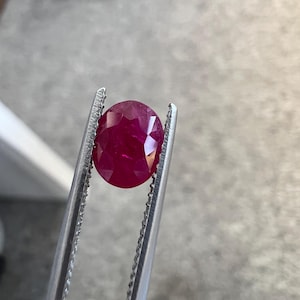 Könnte beinhalten: Ein leuchtender, oval geschliffener Rubin mit tiefroter Farbe und facettierter Oberfläche, der sicher von einer silberfarbenen Metallpinzette gehalten wird. Der Edelstein zeigt eine klare Lichtreflexion auf seiner polierten Oberfläche.