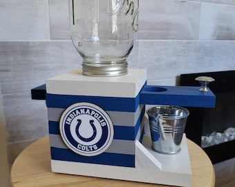 May include: A wooden Indianapolis Colts-themed water dispenser with a large glass mason jar on top. The dispenser is painted white, blue, and gray, with a small metal bucket and a Colts logo. It sits on a wooden stool.