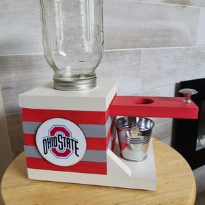 May include: Wooden dispenser with a clear glass jar, a red, white, and gray striped base, and a small metal cup. The base features the Ohio State logo.