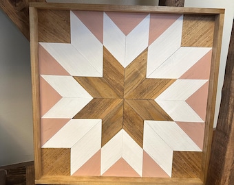 Puede incluir: Una pieza de arte de pared enmarcada en madera con un diseño de estrella geométrica rosa y blanca. La estrella está hecha de madera y pintada en un esquema de color rosa claro y blanco.