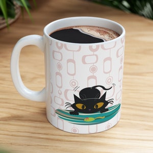 May include: White ceramic coffee mug featuring a black cat design. The cat is on a blue and green surfboard with a yellow fish. The mug has a pink and white geometric pattern.
