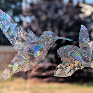 May include: Two iridescent hummingbird silhouettes with a stained glass design. The hummingbirds are facing opposite directions and are made of a clear material with a rainbow sheen.