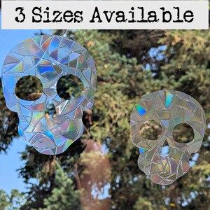 May include: Two iridescent skull window clings with a mosaic design. The text "3 Sizes Available" is at the top. The skulls are transparent and reflect light, creating a rainbow effect. Perfect for Halloween or gothic decor.
