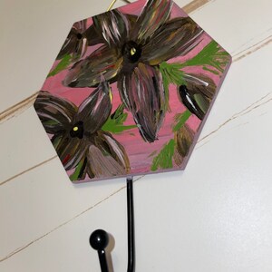 May include: A hexagonal wooden wall hook with a pink background and painted floral design. The hook is black metal and has a rounded end.