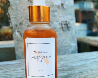 May include: A clear glass bottle with a gold cap and a white label. The label says "Nine Hills Farm Calendula Oil" and "1 fl oz".